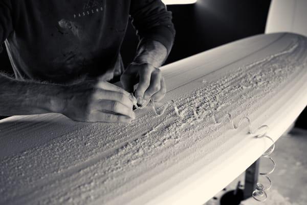 Ollie Cooper planing down the stringer whilst shaping a surfboard. Black and white photo.