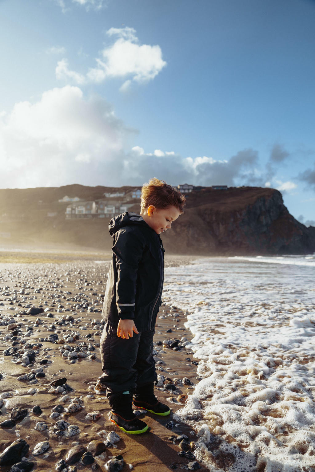 A boy in a black coat standing at a pebble strewn shoreline with the sun at his back