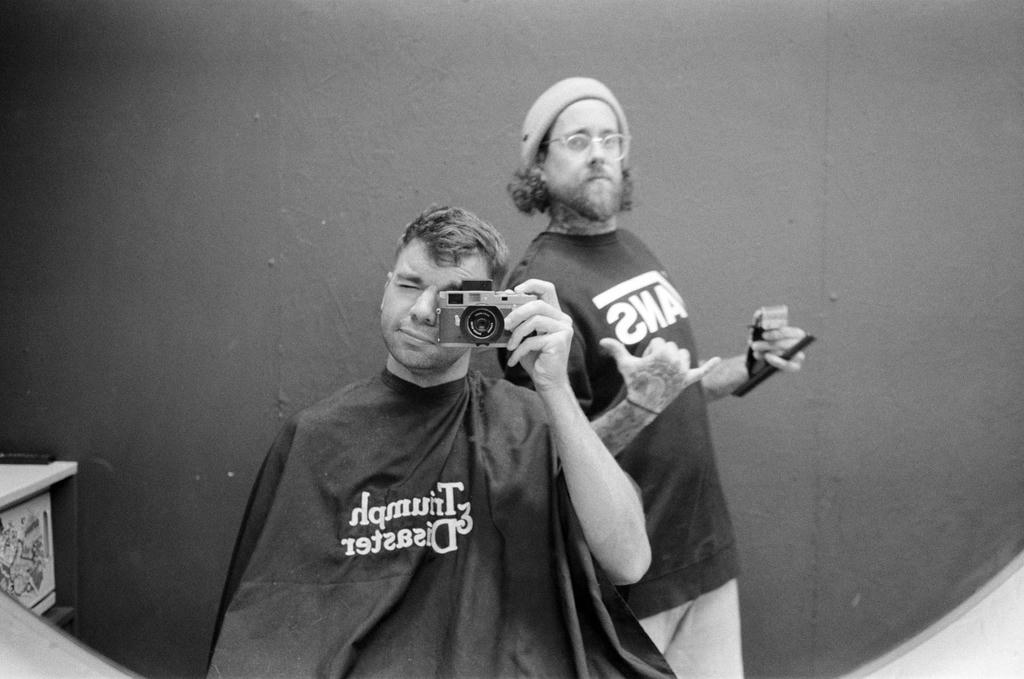 Jamie Dumont holding a Leica M2, taking a picture of himself and barber Aaron Brewer in a mirror.